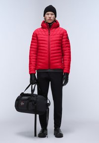 Rød pufferjakke med hætte, kviltet design, båret over en sort sweater. Sorte handsker og bukser, holder en sort sportstaske med logo detalje.