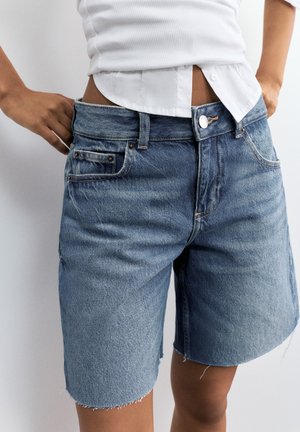 Persona che indossa pantaloncini di jeans blu a vita alta con orli grezzi e una canotta bianca a coste sopra una camicia bianca con bottoni.