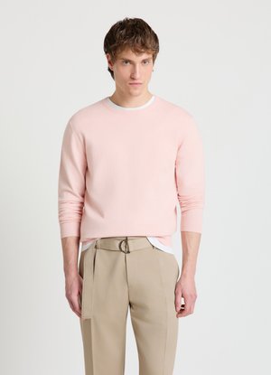 Uomo che indossa un maglione rosa chiaro sopra una camicia bianca e pantaloni beige a vita alta con cintura in tessuto, in piedi su uno sfondo chiaro e uniforme.