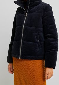 Svart kordflosspufferjacka med silverdragkedja och tryckknappsfickor, bärs över en texturerad orange stickad kjol.