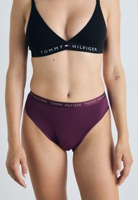 Fekete bralette háromszög alakú és logós pánttal; sötétlila magas derekú fehérneművel párosítva, amely logós derékrésszel és sima textúrával rendelkezik.