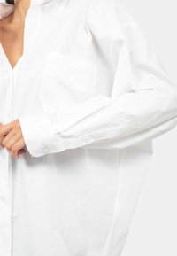 Camicia bianca con bottone, realizzata in tessuto leggero. Presenta una tasca frontale e maniche arrotolate, con una vestibilità rilassata e drappeggiata.