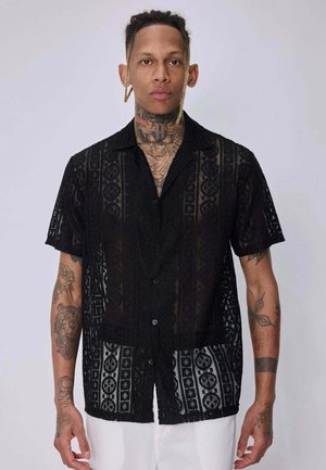 Homme avec des tatouages portant une chemise noire à manches courtes en dentelle à motifs et un pantalon blanc, debout devant un fond clair uni.