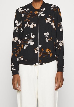 Veste noire à fermeture éclair avec imprimé floral blanc et marron, portée avec un pantalon blanc, visible des épaules aux hanches.