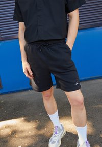 Svarta sportshorts med elastiskt midjeband och snörning, med en vit logga som accent. Matcher med vita strumpor och grå sneakers.