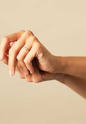 Zwei Hände, die miteinander verschränkt sind, wobei eine Hand einen Goldring mit einem kleinen Diamanten am Ringfinger trägt, vor einem schlichten beigen Hintergrund.