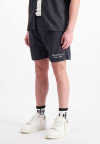 Zwarte korte broek met een patroon en een gestructureerd design, voorzien van witte geborduurde tekst. Gecombineerd met witte sneakers en sokken met zwarte accenten.