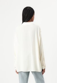Blusa crema a maniche lunghe con un taglio morbido, caratterizzata da un colletto e dettagli con bottoni sulle maniche. Texture liscia.