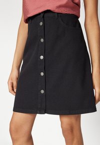 Black button-front denim skirt worn with a red textured top, shown from waist to mid-thigh, with hands relaxed at sides.