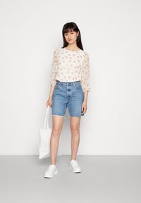 Blusa floral com mangas longas, combinada com calções de ganga com bainha desfiada. A modelo segura uma bolsa de tecido branca e usa sapatilhas brancas.