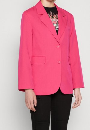 Mujer con una chaqueta rosa brillante de dos botones sobre una camiseta negra con gráfico y pantalones negros, de pie frente a un fondo claro y liso.