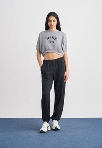 Γκρι κοντό φούτερ με μαύρο λογότυπο "NIKE 1972", συνδυασμένο με μαύρο στενό παντελόνι φόρμας και λευκά αθλητικά παπούτσια με πράσινες λεπτομέρειες.