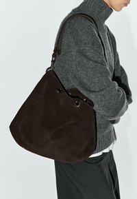 Borsa in camoscio marrone con forma arrotondata, dotata di tracolla superiore e occhielli in metallo. Design minimalista che mette in risalto la texture e l'artigianalità.