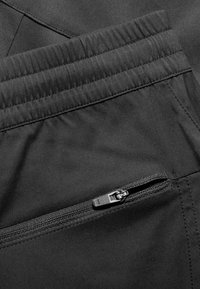 Schwarze Hosen mit elastischem Bund und einer Reißverschlusstasche. Der Stoff wirkt glatt und langlebig und weist kontrastierende Nähte auf.