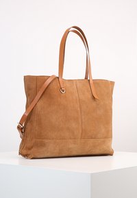 Bruine suede tote bag met twee leren schouderbanden en een afneembare verstelbare crossbodyband, rustend op een witte ondergrond.