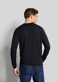 Schwarzer Pullover mit langen Ärmeln, rundem Halsausschnitt und geripptem Saum. Über blauen Jeans getragen, zeigt er einen glatten Stoff und ein einfaches Design.