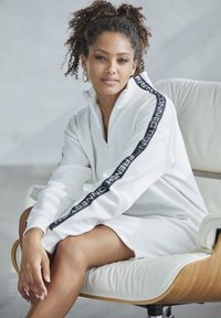 Weißes Zip-Hoodie-Kleid mit schwarzem Branding an den Ärmeln, im lässigen Schnitt und aus weichem, strukturiertem Material.