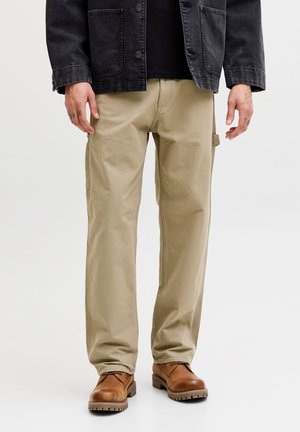 Homme portant un pantalon cargo beige, des bottes marron lacées et une veste noire, debout sur un fond blanc uni.