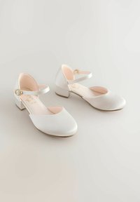 Een paar witte satijnen lage hakken met enkelbandjes en gouden gespen op een lichte achtergrond.