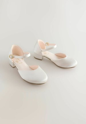 Pár bílých saténových lodiček na nízkém podpatku s kotníkovými pásky a zlatými sponami na světlém pozadí.