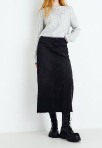 Grauer Strickpullover, langer schwarzer Rock mit Frontschlitz und schwarze Schnürstiefel mit grober Sohle, die einen Lagenlook mit kontrastierenden Texturen erzeugen.