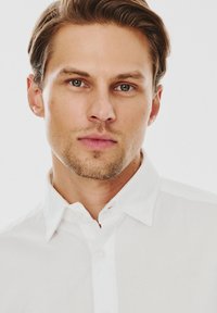 Biała koszula z guzikami i klasycznym kołnierzykiem. Gładka tekstura, brak wzorów. Zbliżenie na twarz mężczyzny z krótkimi włosami i delikatnym zarostem.