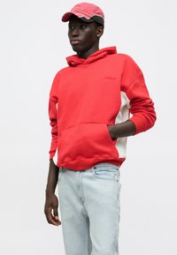 Felpa rossa con tasca a marsupio e pannelli laterali bianchi; abbinata a jeans azzurri chiari. Cappellino rosa con dettagli logori.