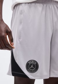 Άνδρας που φοράει ανοιχτόγκρι αθλητικά σορτς Paris Saint-Germain με μαύρη τριγωνική λεπτομέρεια και λογότυπο κοντά στον ποδόγυρο.