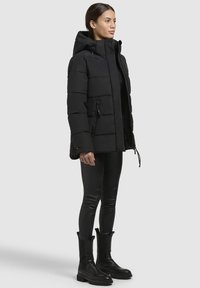 Doudoune noire avec capuche, comportant des panneaux matelassés, des zips sur les côtés et une coupe ajustée, assortie à un legging noir et des bottines.