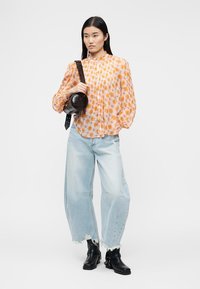 Νεαρή γυναίκα που φοράει πορτοκαλί μπλούζα με λουλούδια, φαρδιά τζιν ανοιχτού χρώματος με ξεφτισμένα τελειώματα, μαύρα μποτάκια αστραγάλου και κρατάει μια στρογγυλή μαύρη δερμάτινη τσάντα.