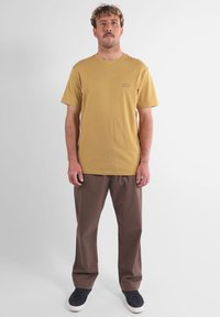 T-shirt en coton jaune à manches courtes avec un petit logo, associé à un pantalon marron ample et des baskets noires. Design simple, sans motifs.