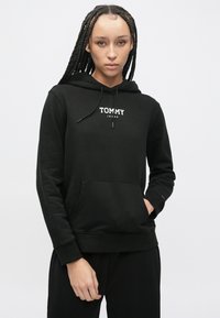 Čierna mikina s dvojitým kapucňou z mäkkej látky, s predným vreckom a šnúrkami na kapucni. Na hrudi je bielym písmom zobrazené logo "TOMMY JEANS".