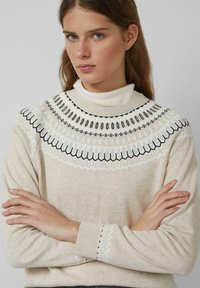 Beżowy sweter z wysokim kołnierzem, zdobiony czarno-białymi geometrycznymi haftami wokół dekoltu. Miękka tekstura i luźny krój.