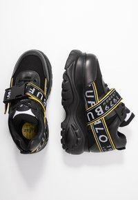 Svarta sneakers med meshoch och syntetiskt övre, gula detaljer, justerbar rem, strukturerad sula och logotypdetaljer på remmen.