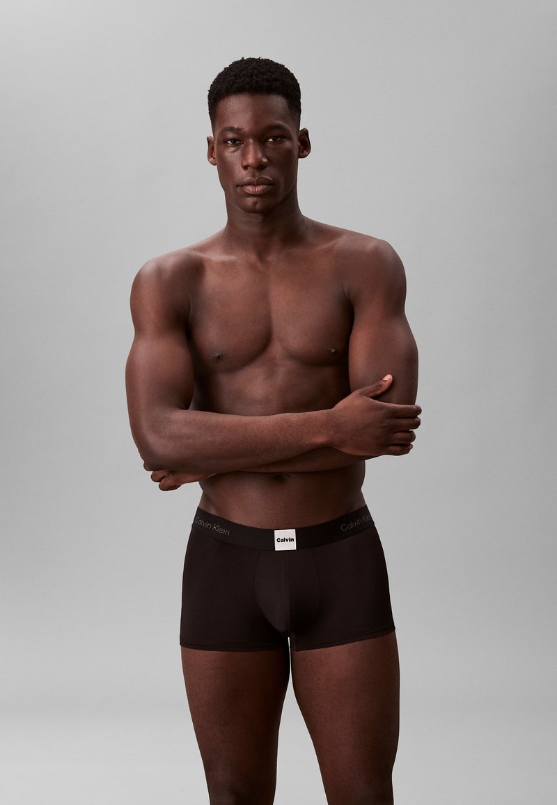 Boxer shorts neri in cotone con una cintura logo Calvin Klein, indossati da un modello con una corporatura muscolosa, su uno sfondo grigio neutro.