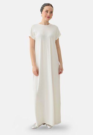 Vrouw draagt een lange, losse witte jurk met korte mouwen, zilveren schoenen en een zwart-wit gestreept hoofddoek, staand op een effen achtergrond.