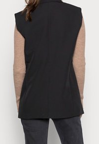 Black sleeveless blazer with a structured design, featuring a collar, smooth texture, and a tailored fit, worn over a light brown long-sleeve top.