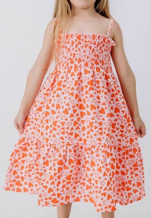 Junges Mädchen trägt ein ärmelloses rosa Kleid mit orangefarbenen Herzmustern und hält die Seiten des Kleides vor einem schlichten weißen Hintergrund fest.