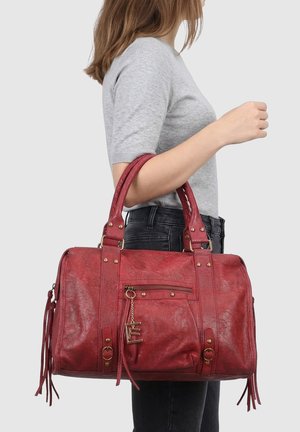 Femme portant un haut gris à manches courtes et un jean noir, tenant un grand sac à main en cuir rouge avec une poche zippée et un porte-bonheur doré.
