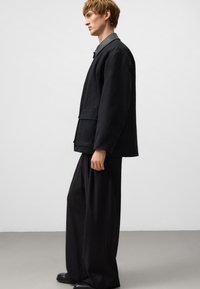 Czarna, oversize kurtka, wykonana z bawełny, luźny krój, spiczasty kołnierz, kieszeń na piersi, zestawiona z szerokimi czarnymi spodniami i czarnymi butami.