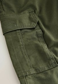 Olijfgroen stof met een gladde textuur heeft een klepzak en contrasterende stiksels, wat het ontwerp van cargo pantalons benadrukt.