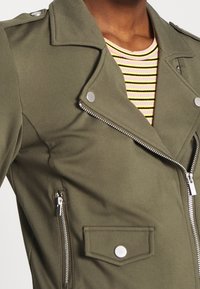 Close-up of a person wearing an olive green biker jacket with silver zippers and buttons over a pink, yellow, and black striped ribbed shirt.