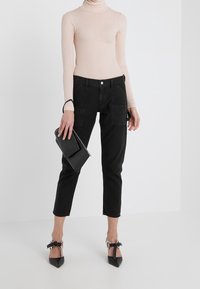 Jeans de talle alto negros con bolsillos frontales, un suéter de cuello alto ajustado de color rosa claro y zapatos negros de punta con detalles de lazo. Sosteniendo un clutch negro.
