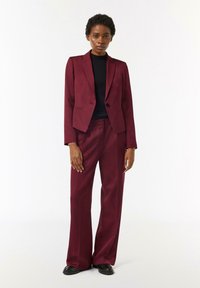 Burgundowa marynarka w fasonie dopasowanym z pojedynczym guzikiem, w zestawie z szerokimi spodniami. Materiał wydaje się gładki; nałożona na czarną bluzkę.