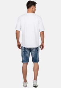 Homme se tenant de dos, portant un T-shirt blanc uni, un short en denim bleu retroussé et des baskets blanches avec des chaussettes blanches courtes.