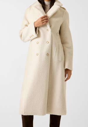 Femme portant un long manteau en laine crème avec grand col, boutons croisés et ceinture latérale, associé à un pantalon foncé.