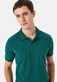 Turkos polo-shirt med en texturerad krage och korta ärmar. Har två knappar vid halslinningen och en figurnära design.
