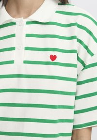 Weißes Poloshirt mit grünen Horizontalstreifen, mit einer roten Herzstickerei auf der linken Seite. Hergestellt aus weichem Baumwollmaterial.