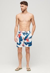 Bañadores de hombre en tejido blanco con estampados de hojas tropicales en azul y rojo. Cuentan con una cinturilla elástica con cordón y dos bolsillos laterales.