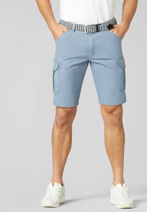 Mann trägt hellblaue Cargo-Shorts mit Vorder- und Seitentaschen, einen geflochtenen Gürtel, weiße Sneakers und ein weißes T-Shirt, steht mit den Händen in den Taschen.
